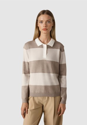 Kobieta w beżowym i białym długim swetrze polo w szerokie paski, beżowych spodniach, stojąca na tle jednolitego jasnoszarego tła.