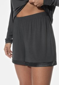 Zwarte shorts met een gladde satijnen zoom, een elastische tailleband en zachte stof, weergegeven met een hand die het bovenste kledingstuk aanpast.