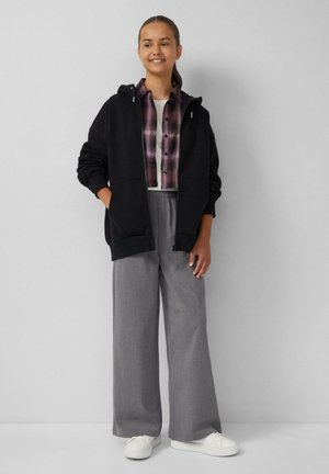 Zwarte hoodie met rits, gedragen over een geblokt overhemd met knopen. Grijze wijde broek en witte sneakers maken de outfit compleet.