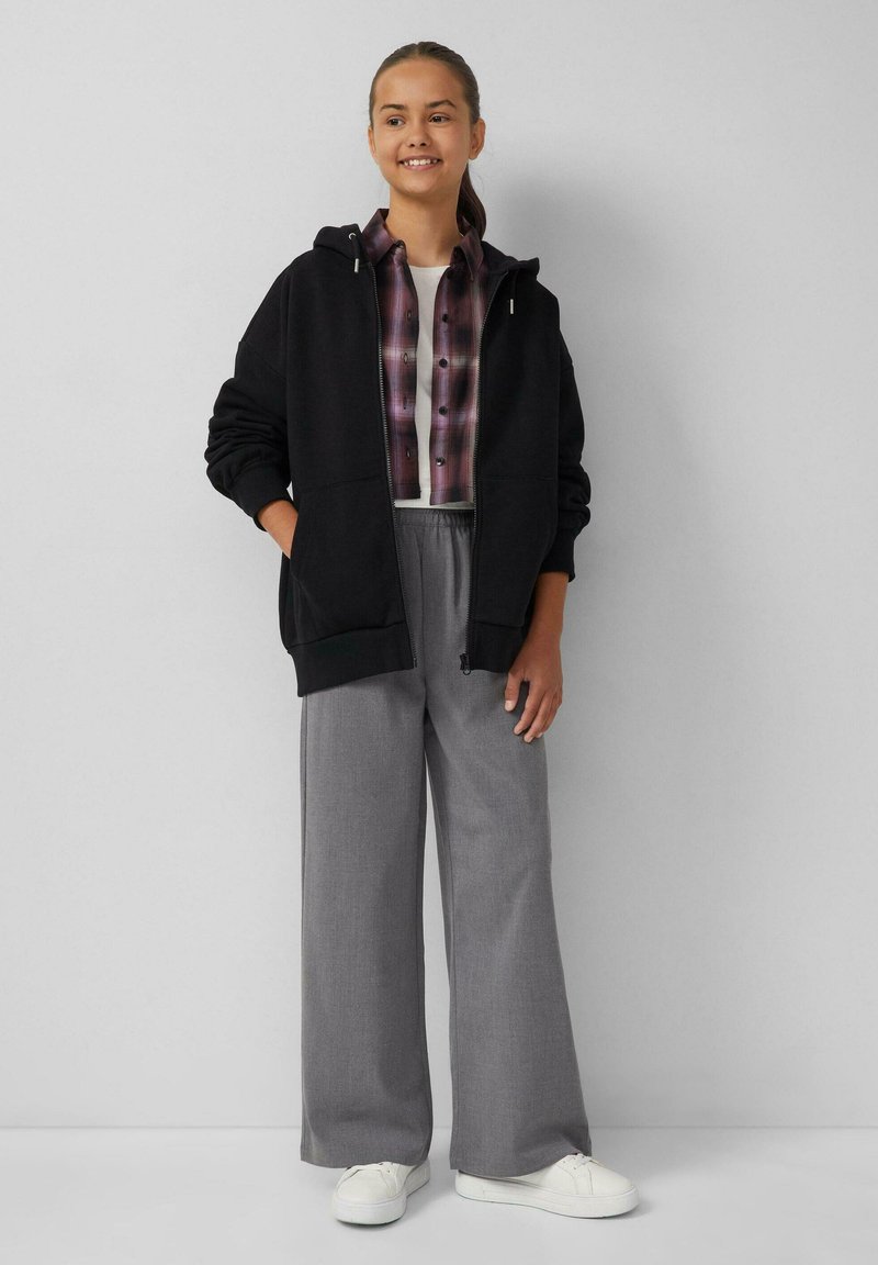 Zwarte hoodie met rits, gedragen over een geblokt overhemd met knopen. Grijze wijde broek en witte sneakers maken de outfit compleet.