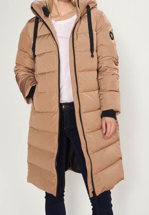 Kobieta ubrana w długi beżowy płaszcz puchowy z czarnym zamkiem błyskawicznym i troczkami, na białej koszuli i ciemnoniebieskich dżinsach.