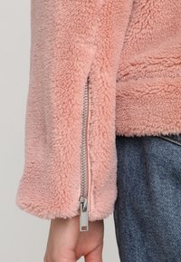Różowa polarowa kurtka o teksturowanej powierzchni, z metalowym zamkiem błyskawicznym przy mankiecie i miękkim, pluszowym materiałem. Prosty, casualowy design bez wzor�ów.