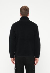 Black fleece jacket with a high collar, textured fabric, and relaxed fit. Visible stitching and no prominent hardware details.