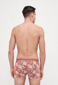 Pantaloni boxer con una base rosso ruggine e un motivo floreale bianco. Presentano un elastico a righe di contrasto in vita. Il materiale sembra morbido ed elastico.