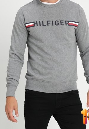 Mężczyzna noszący szarą bluzę z nadrukiem "HILFIGER" i czerwono-biało-niebieskimi paskami na klatce piersiowej, w połączeniu z czarnymi spodniami.