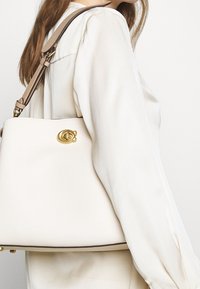 Bílá kožená kabelka se strukturovaným tvarem, zlatými kovovými prvky a kontrastním hnědým popruhem. Disponuje logem jako akcentem a hladkým povrchem.