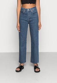 Jeans en denim bleu taille haute avec une coupe droite et une texture légèrement décolorée, associés à des sandales noires.