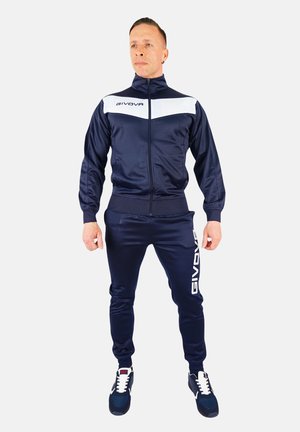 Navy tracksuit made of smooth material, featuring a white stripe, zip-up jacket, and Givova branding on the pants. Black and white sneakers.