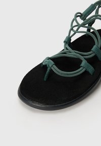 Sandal cu fermoar negru din suede, cu bretele elastice de culoare teal, având o talpă plată și un design complex de șireți pe partea superioară.