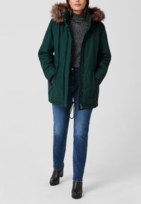 Parka verte avec capuche en fausse fourrure, dotée d'une fermeture éclair à l'avant, d'une taille ajustable par cordon et de poches latérales, portée avec un jean bleu et des bottes noires.