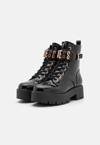 Bottines en cuir verni noir avec des semelles épaisses et texturées, des accessoires en ton doré, et une sangle proéminente portant le mot "Guess."