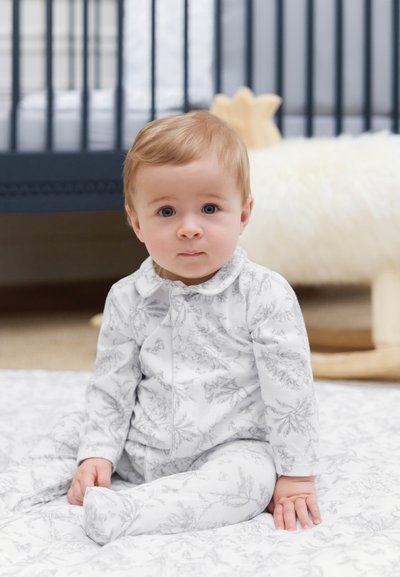 Λευκή πιτζάμα με λεπτό γκρι σχέδιο φύλλων, με γιακά και άνετη εφαρμογή, σε καθισμένο παιδί με ανοιχτά καστανά μαλλιά και μπλε μάτια.