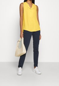 Donna che indossa un top giallo senza maniche, pantaloni blu navy, sneakers bianche e tiene una borsa beige su uno sfondo bianco uniforme.