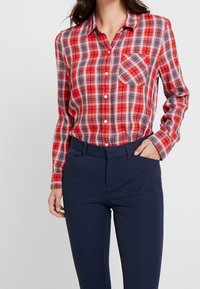 Chemise à carreaux rouge et noir à boutons, manches longues et une poche poitrine, associée à un pantalon ajusté bleu marine foncé. Texture de tissu lisse.