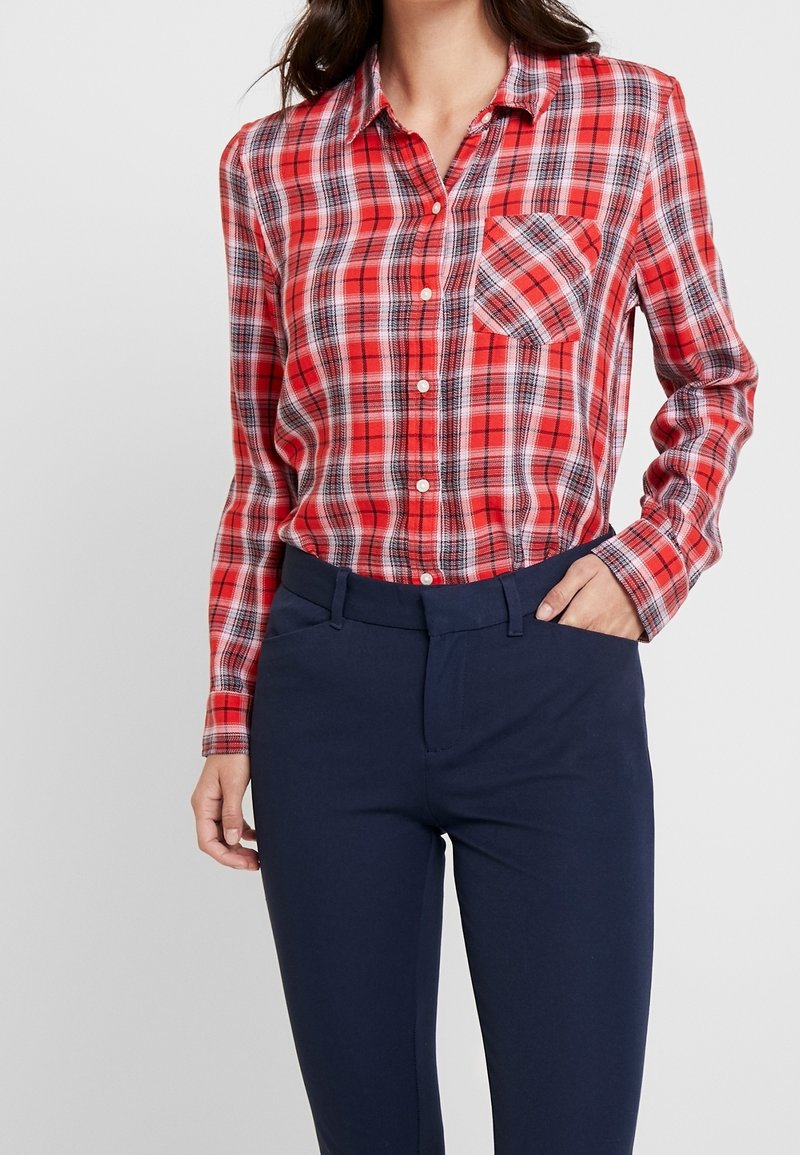 Chemise à carreaux rouge et noir à boutons, manches longues et une poche poitrine, associée à un pantalon ajusté bleu marine foncé. Texture de tissu lisse.