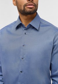 Blauwe textuurshirt met een subtiel patroon, voorzien van een puntkraag en donkere knopen. De stof lijkt glad met een gestructureerde pasvorm.