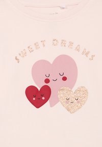 Trih nasmiješenih srca sa zatvorenim očima, jedan ružičast, jedan crven, jedan sjajni zlatni, ispod sjajnog teksta "SLATKI SNOVI" na svijetloj tkanini.