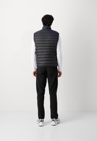 Hombre de pie de espaldas, vistiendo un chaleco acolchado negro, una camisa de manga larga blanca, pantalones negros y zapatillas deportivas blancas, con un fondo liso.