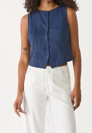 Femme portant un haut sans manches bleu marine à boutons et un pantalon blanc taille haute avec un cordon à la taille, debout devant un fond uni.