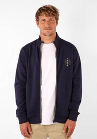 Sweatshirt zippé marine en tissu doux, avec un col haut, des poignets côtelés et un design brodé sur la poitrine gauche.