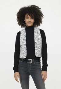 Strickweste im weißen und grauen Fischgrätmuster, getragen über einem schwarzen Rollkragenpulli, kombiniert mit dunklen Jeans und einem dekorativen Gürtel.