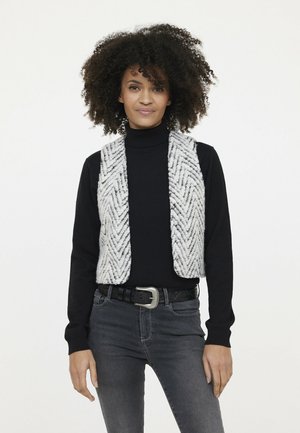 Gehaakte vest in een wit en grijs visgraatpatroon, gedragen over een zwarte coltrui, gecombineerd met donkere jeans en een decoratieve riem.