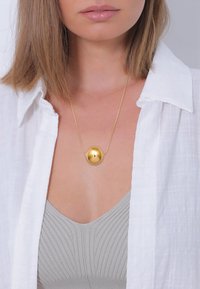 Gul pendantsmykke med oval form, hængende p�å en fin kæde. Modellen bærer en hvid skjorte over en grå ribbet top.