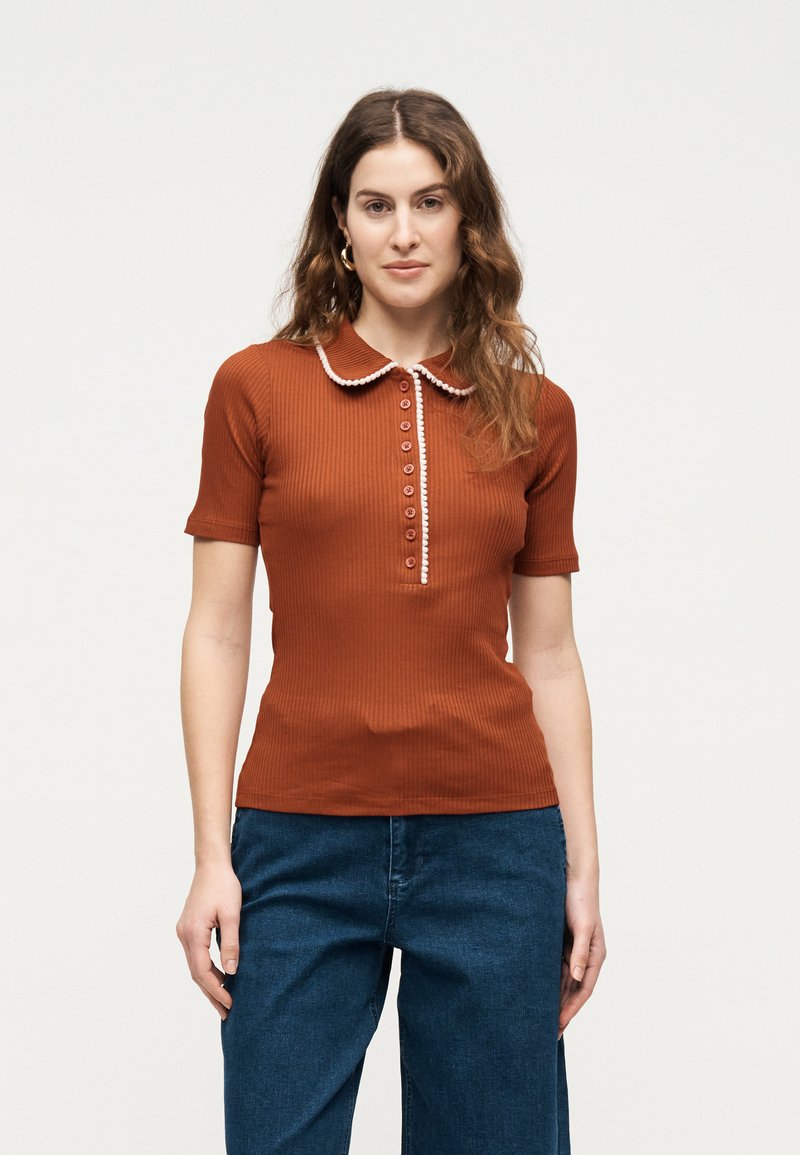 Γυναίκα με κυματιστά καστανά μαλλιά, που φοράει κοντομάνικο πόλο σε σκουρόχρωμο πορτοκαλί με ριγέ υφή, λευκή επένδυση και κουμπιά, συνδυασμένο με μπλε τζιν.