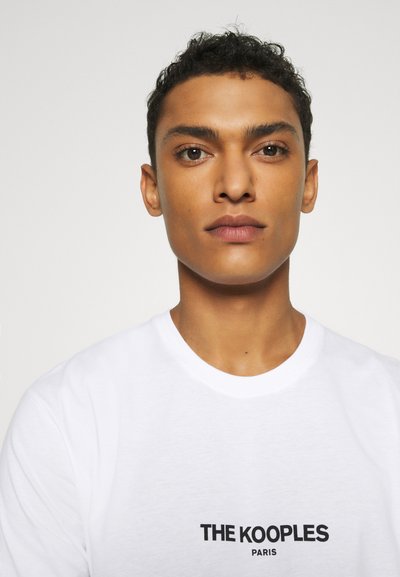 Tricou din bumbac alb, cu guler rotund și mâneci scurte, având inscripția "THE KOOPLES PARIS" imprimate cu litere mari de culoare neagră pe față.