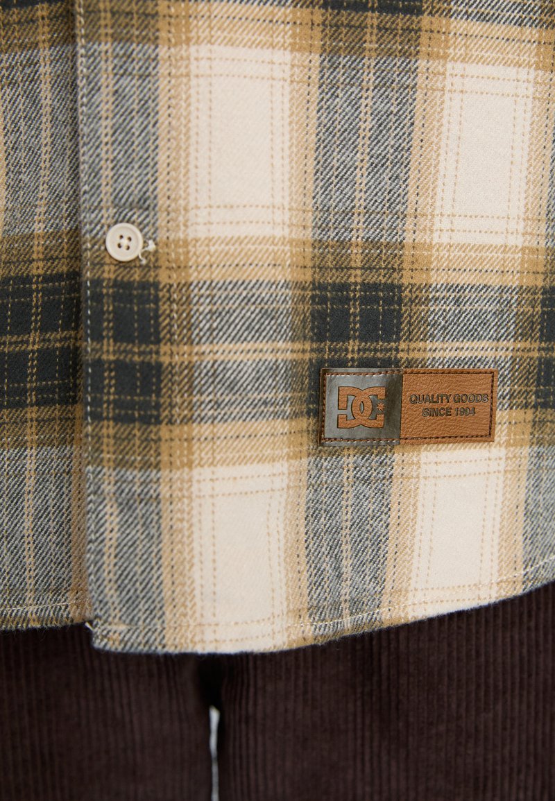 Koszula flanelowa w kratę w odcieniach beżu, brązu i granatu z zapięciem na guziki. Posiada skórzaną metkę marki DC. Miękka tekstura i standardowy krój kołnierza.
