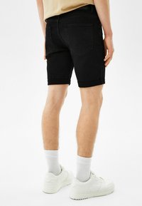 Mies, który ma na sobie czarne dżinsowe szorty z mankietami, białe skarpetki i białe tenisówki, stoi na tle białym, zwrócony plecami.