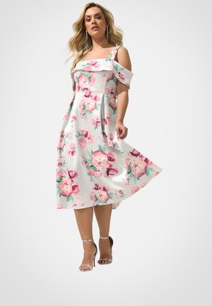 Kvinde iført en hvid midi-kjole med pink blomsterprint og kolde skulderstropper, kombineret med sølvfarvede høje hæle sandaler.