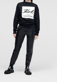 Sudadera negra con logo rectangular blanco, pantalones de cuero negros y zapatos de plataforma negros gruesos con detalle de cadena plateada.