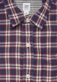 Chemise à carreaux rouge, blanche et bleue à boutons. Présente un col classique, des boutons blancs, une poche sur le côté gauche, et une texture de tissu tissé doux.