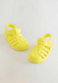 Gelbe Kunststoffsandalen mit geschlossener Zehe, verstellbarem Riemen und durchbrochenen Designs zur Belüftung. Leichte und flexible Bauweise.