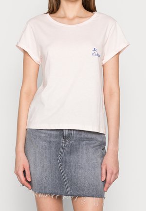Femme portant un T-shirt rose clair à manches retroussées avec le texte « Je t’aime » et une mini-jupe en jean gris asymétrique à bords effilochés.