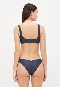 Tamni sivi bikini set s gornjim dijelom s izrezom na leđima, širokim trakama i donjim dijelom s minimalnom pokrivenošću. Prisutna je glatka tekstura i klasični dizajnerski elementi.