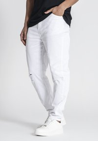 Jeans de denim blanco con un corte relajado, que presentan detalles desgastados en las rodillas. Combinados con zapatillas blancas y una camisa negra.