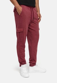 Herren Jogginghose in Kastanienrot aus weichem Stoff, mit seitlichen Taschen und gerippten Bündchen, kombiniert mit weißen Sneakern.