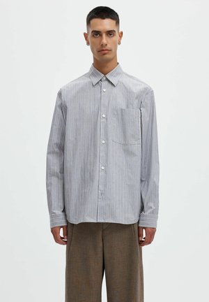 Jeune homme aux cheveux courts avec des boucles d'oreilles, portant une chemise boutonnée à rayures verticales grises et blanches et un pantalon marron à jambes larges, se tenant devant un fond uni.