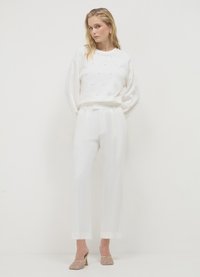 Sweat-shirt blanc texturé avec des accents décoratifs, associé à un pantalon blanc sur mesure avec un ourlet retroussé. Le modèle porte des chaussures à talons dorés.