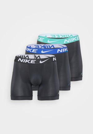 Boxers ajustados de Nike en gris con una cinturilla turquesa, que presentan un ajuste ceñido y una textura suave. El modelo está descalzo, con los brazos relajados a los lados.
