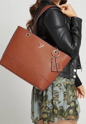 Frau trägt schwarze Lederjacke und geblümten Rock, hält große braune Guess-Tasche mit Markenanhängern über der Schulter.