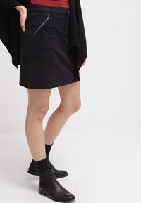 Schwarzer Rock mit einem taillierten Design, der eine seitliche Reißverschlusstasche aufweist. Kombiniert mit schwarzen Stiefeletten und schwarzen Socken. Glatte Textur, knielang.