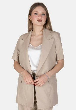 Giovane donna con i capelli lunghi che indossa un blazer beige a maniche corte, una maglietta bianca e pantaloni beige, in piedi davanti a uno sfondo neutro.