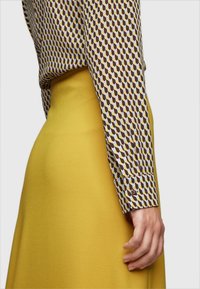 Chemise à manches longues avec un motif géométrique marron et blanc, associée à une jupe jaune unie. La jupe présente une finition lisse et texturée.
