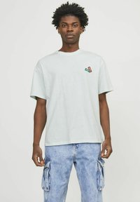 Camiseta de algodón gris claro con cuello redondo y mangas cortas, que presenta un pequeño gráfico en verde y rojo en el lado izquierdo del pecho.