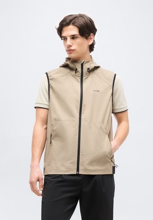 Jonge man met een beige mouwloos hooded vest met zwarte ritssluiting en zijzakken, gecombineerd met een beige shirt met korte mouwen en zwarte broek.
