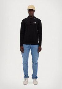 Sort langermet poloshirt med hvite detaljer, kombinert med lyseblå jeans og beige caps. Beige joggesko fullfører antrekket.