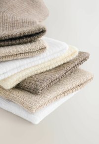 Stapels opgevouwen gebreide wollen truien in neutrale tinten van beige, wit en lichtbruin op een lichte achtergrond.